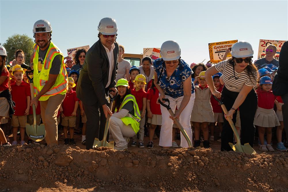 People dressed in hard hats and safety vests use shovels to dig into the ground